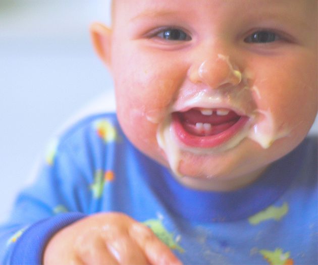 A happy baby wearing a blue shirt, with food smeared on their face and a joyful smile.