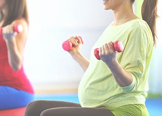 Pregnant mother sitting cross-legged in a prenatal barre lifting small dumbbells