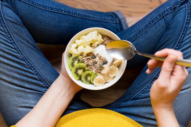 A pregnant woman sat cross legged in blue jeans, holding a bowl of yoghurt, granola and fruit