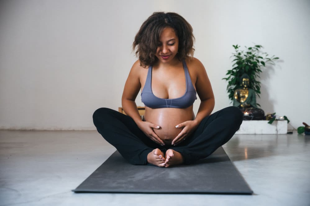A 28 weeks pregnant woman doing yoga on a black yoga mat in room with a white background