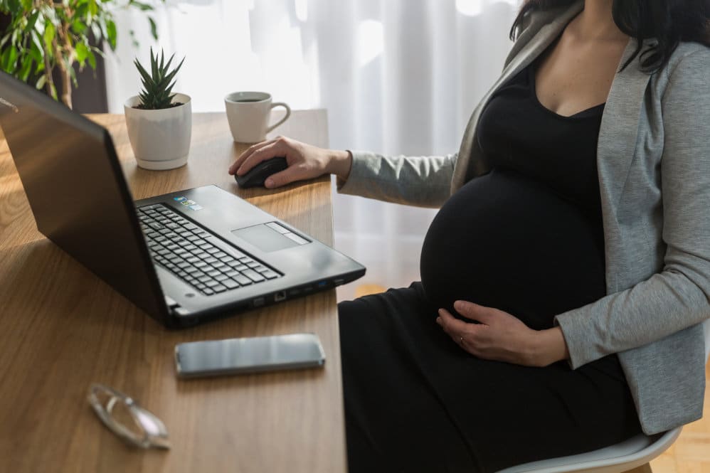 A woman who is 29 weeks pregnant sat at a desk looking at her laptop, holding onto her belly 