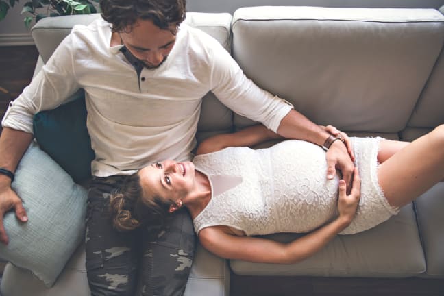 A couple on a sofa, the pregnant woman lying on the mans lap both with their hands on her belly