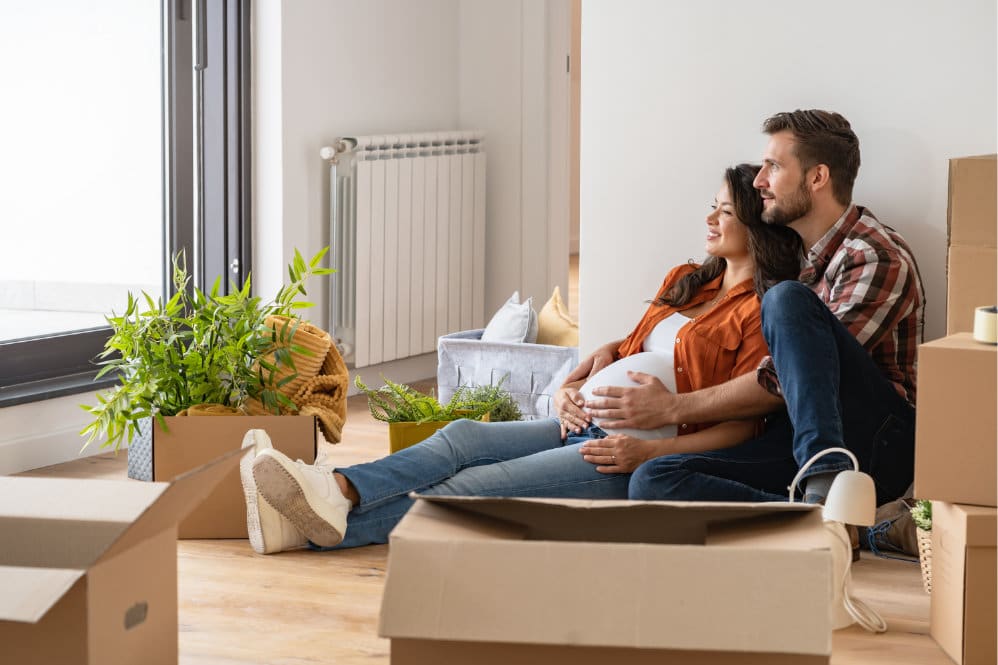 A couple sat in a room surrounded by cardboard boxes while holding on to her baby bump