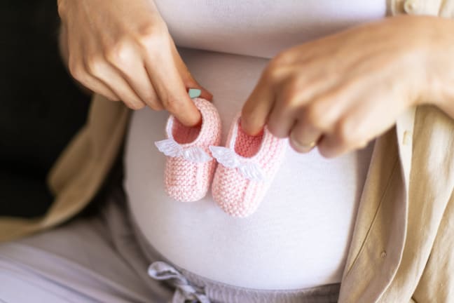 A pregnant woman in a white top and linen shirt holding a pair of pink knitted, newborn booties