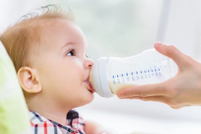 Close-up of a young child drinking milk from a baby bottle held by an adult hand, with measurement markings visible on the bo