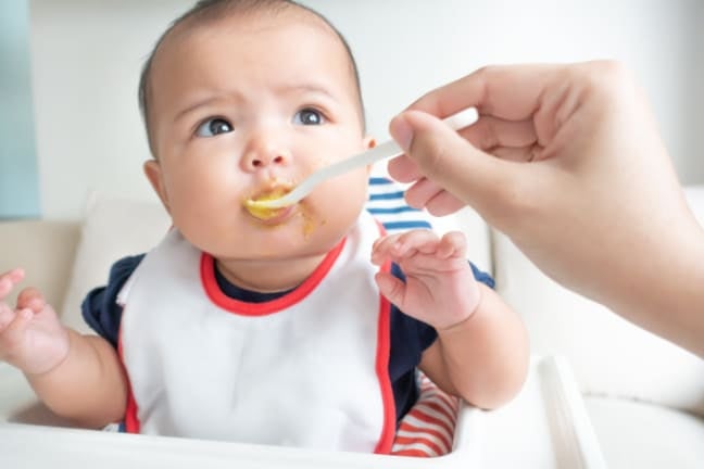 An adult hand offering a spoon of puréed food to a baby during